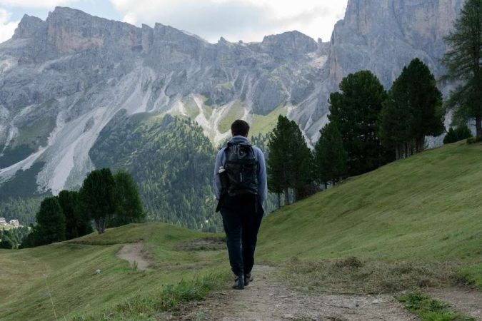 A solo trekker with a black backpack walking toward dramatic rocky Dolomite peaks on a green mountain trail.